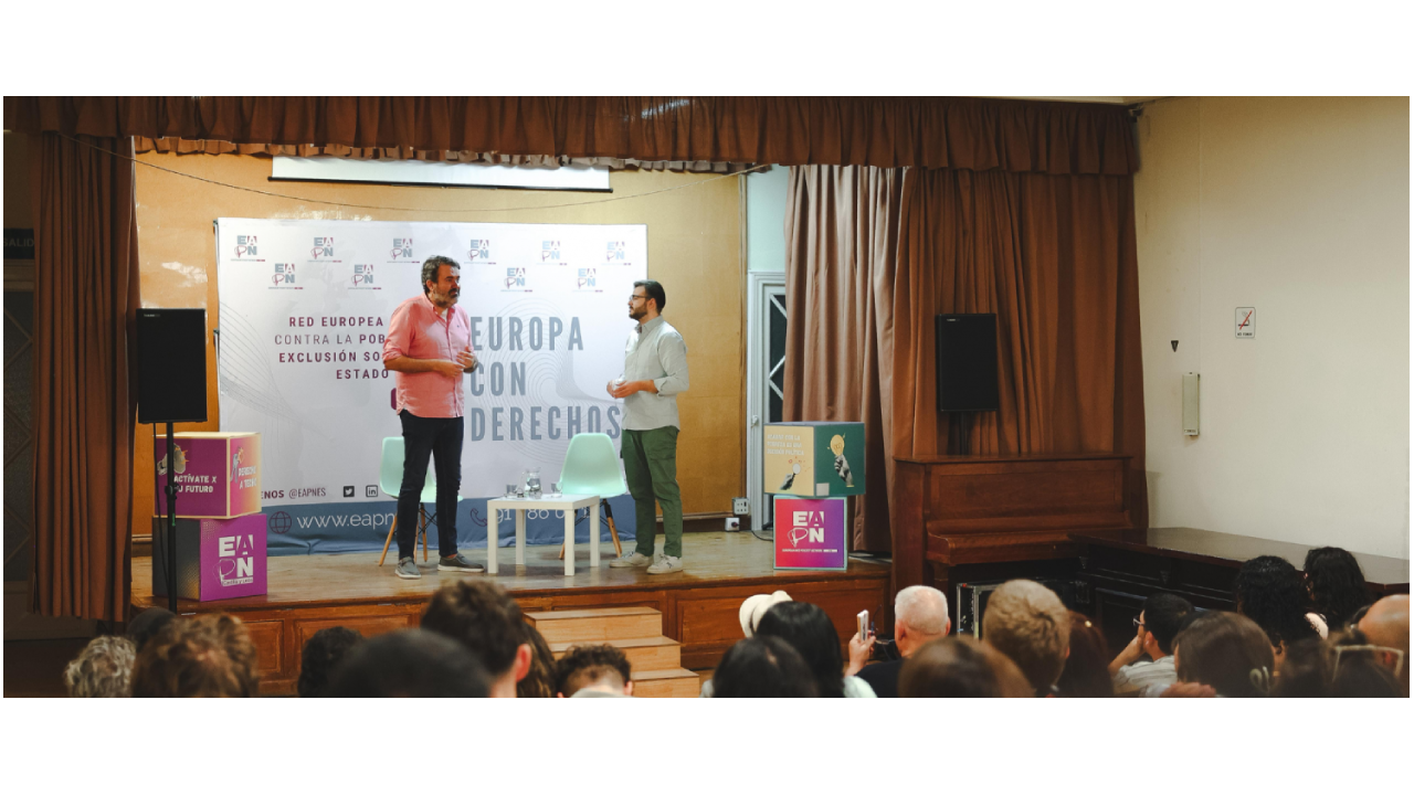 Dos hombres de pie hablan en encima de un escenario con un photocall de EAPN