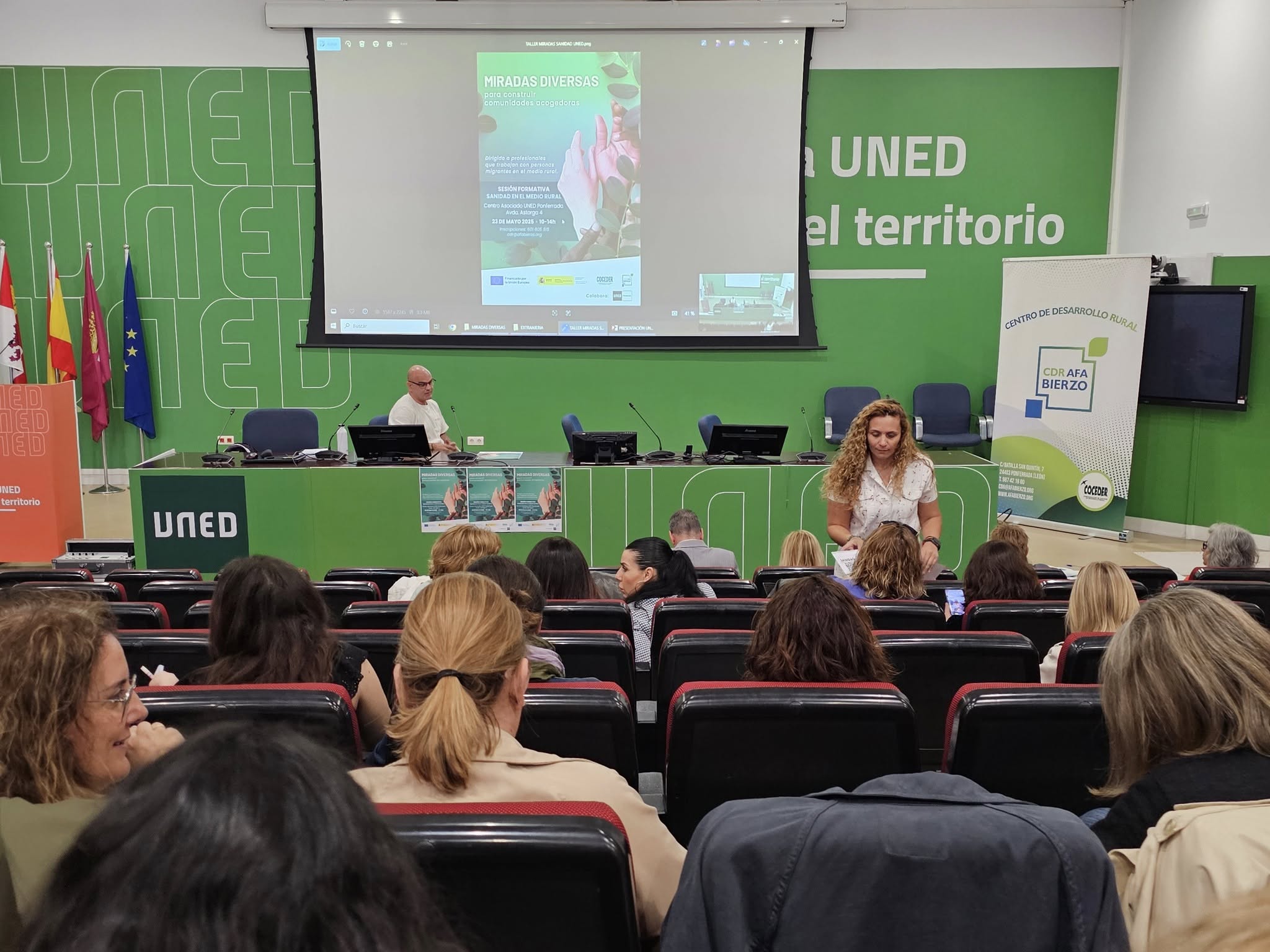 Vista de público escuchando en un salón de actos con una pantalla con un cartel "Miradas diversas para construir comunidades acogedoras "Miradas diversas para construir comunidades acogedoras"