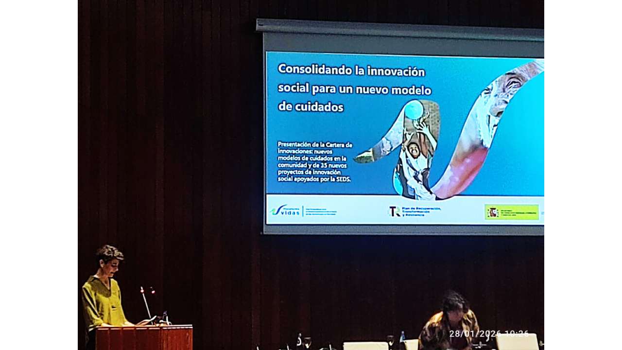 Una persona de pie habla en un atril con una presentación detrás en una pantallla