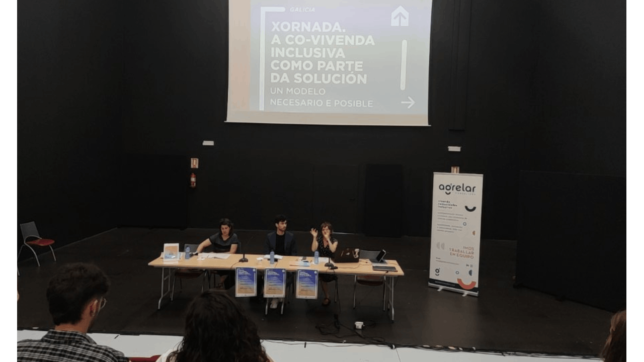 Unas personas sentadas en una mesa hablan delante de una pantalla con un cartel que se lee Jornada A Covivienda inclusiva como parte de la solución