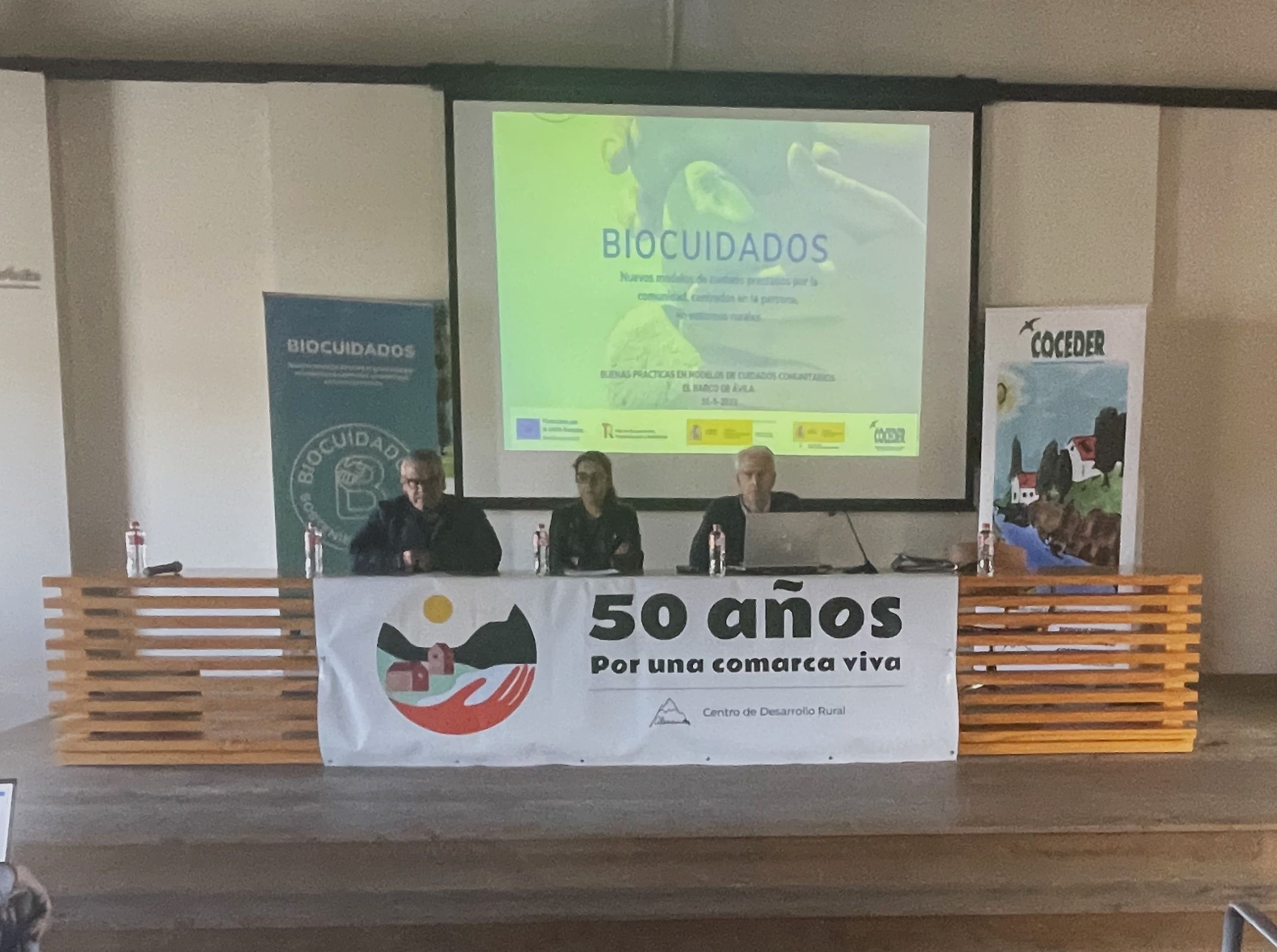 La presidenta de Coceder, el director del CDR de Almazor y un representante del ayuntamiento explican la jornada con una imagen poryectada en el fondo de  Buenas Prácticas de Biocuidados