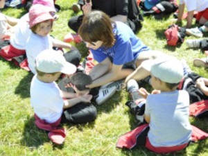 Grupo de niños sentados en una pradera con una monitora enseñando un animal en una mano a uno de los niños